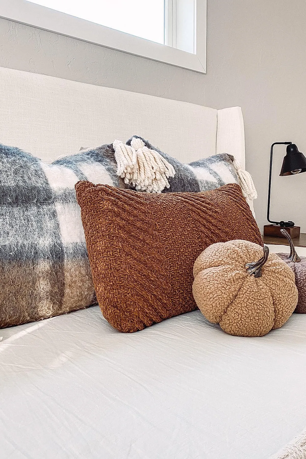 bed with white headboard featuring burnt orange knit rectangular pillow, gray plaid pillows with white tassels, and tan faux shearling pumpkin on white bedding