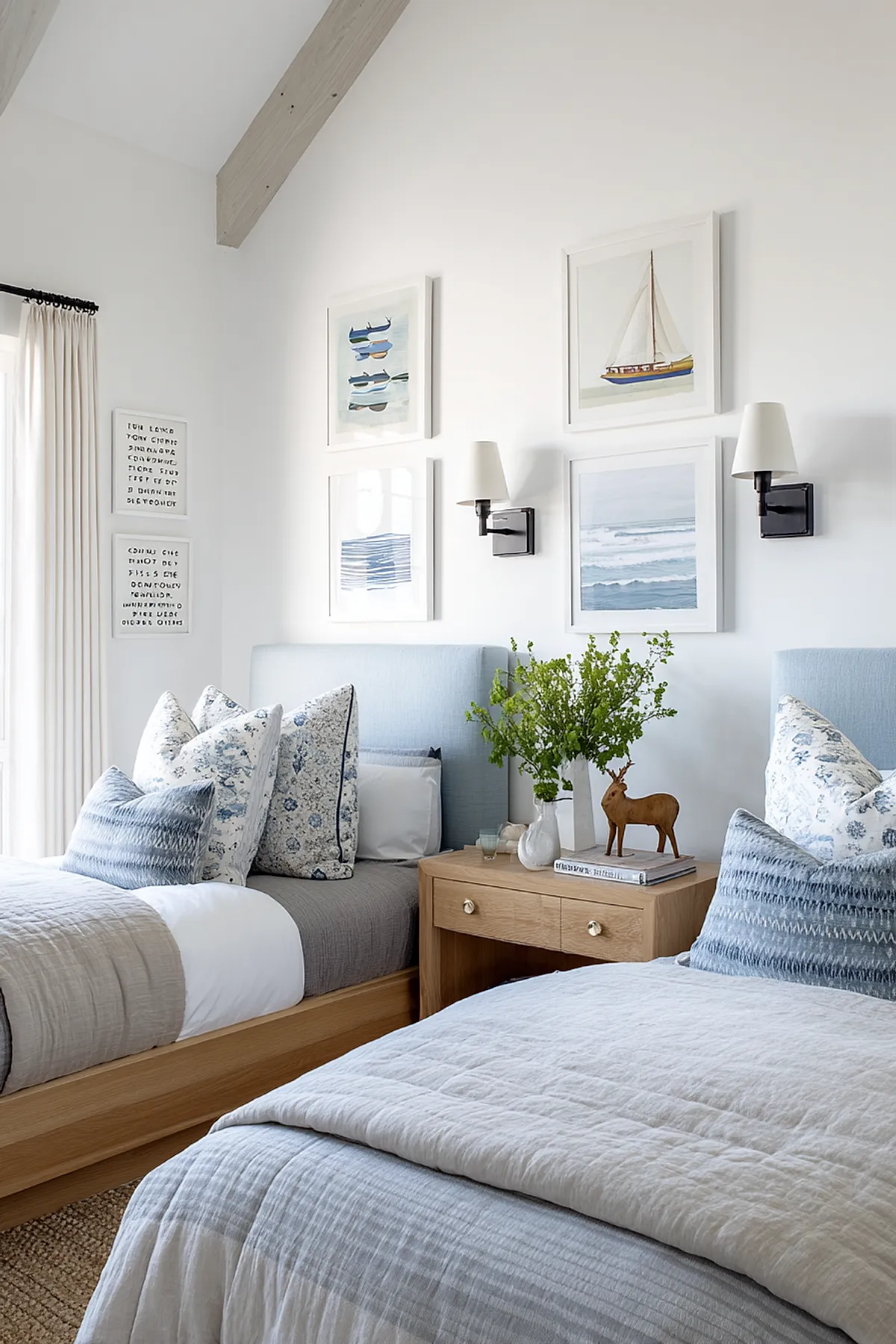 Shared boys bedroom featuring two twin beds with light blue upholstered headboards, gray bedding with patterned pillows, a wooden nightstand with green foliage and decor in center, white walls adorned with framed boat and ocean prints, and two black wall sconces with white shades.